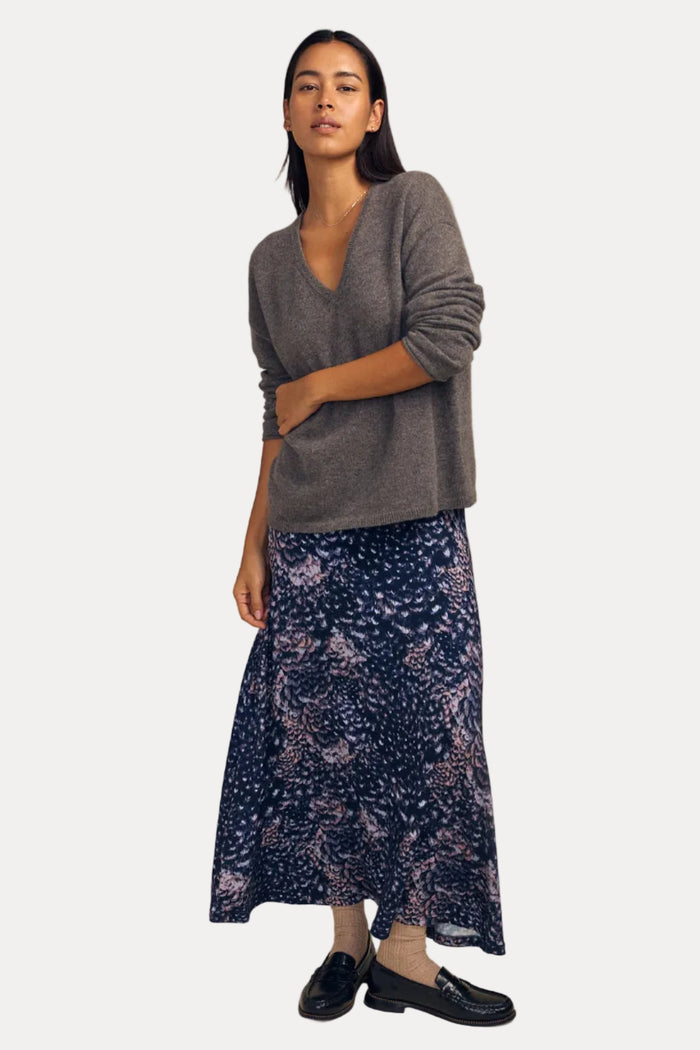 A woman stands against a plain beige studio background wearing a gray V-neck sweater, a navy floral midi skirt, beige socks, and black loafers.