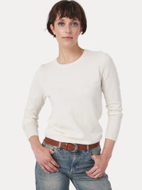A woman modeling a cream-colored, long-sleeve crewneck knit sweater paired with blue jeans and a brown leather belt, shot against a plain light gray studio background.