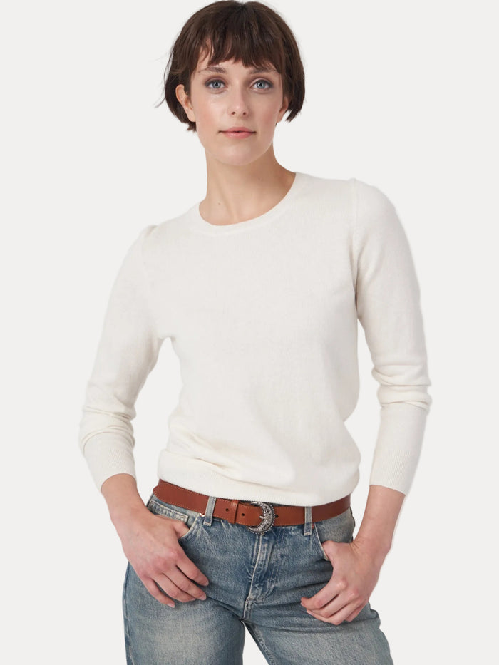 A woman modeling a cream-colored, long-sleeve crewneck knit sweater paired with blue jeans and a brown leather belt, shot against a plain light gray studio background.