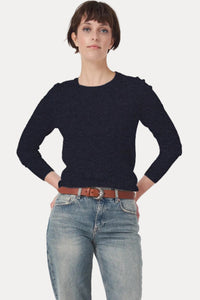 A model stands against a light gray studio background wearing a dark navy long-sleeve knit top, light-wash blue jeans with rolled cuffs, a brown belt, and dark brown shoes.