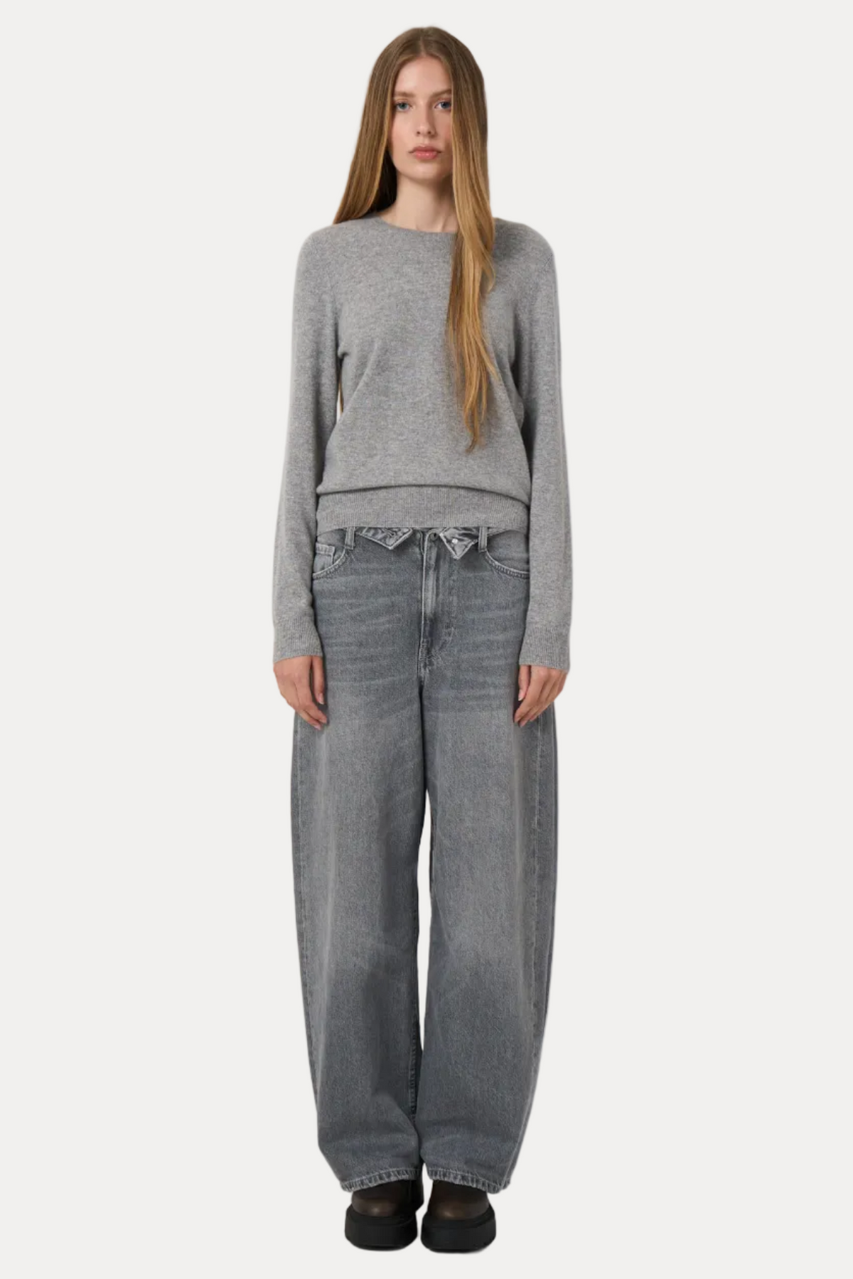 A model stands against a plain white studio background wearing a light gray knit sweater, wide-leg gray jeans, and black shoes.