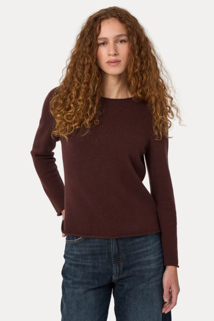 A woman with long, curly light brown hair wearing a dark brown long-sleeve knit sweater and dark blue jeans, standing against a plain light gray studio background.