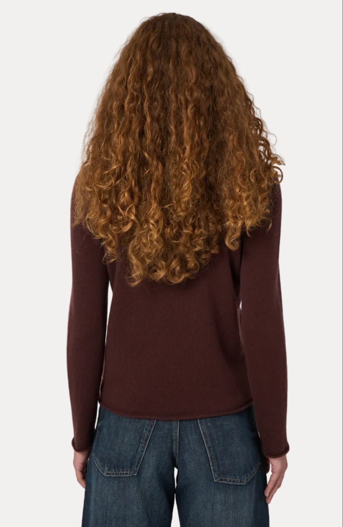 A person viewed from the back wearing a long-sleeve burgundy knit top and blue jeans, with long curly brown hair, against a plain light gray background.