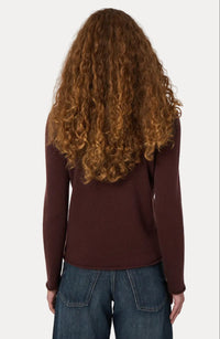 A person viewed from the back wearing a long-sleeve burgundy knit top and blue jeans, with long curly brown hair, against a plain light gray background.