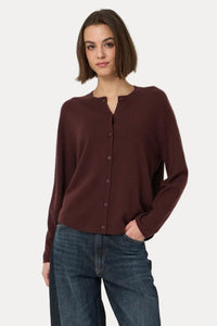 A woman modeling a dark brown long-sleeve button-front cardigan top with dark blue wide-leg jeans and brown leather boots, standing against a plain light gray studio background.