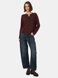 A woman modeling a dark brown long-sleeve button-front cardigan top with dark blue wide-leg jeans and brown leather boots, standing against a plain light gray studio background.