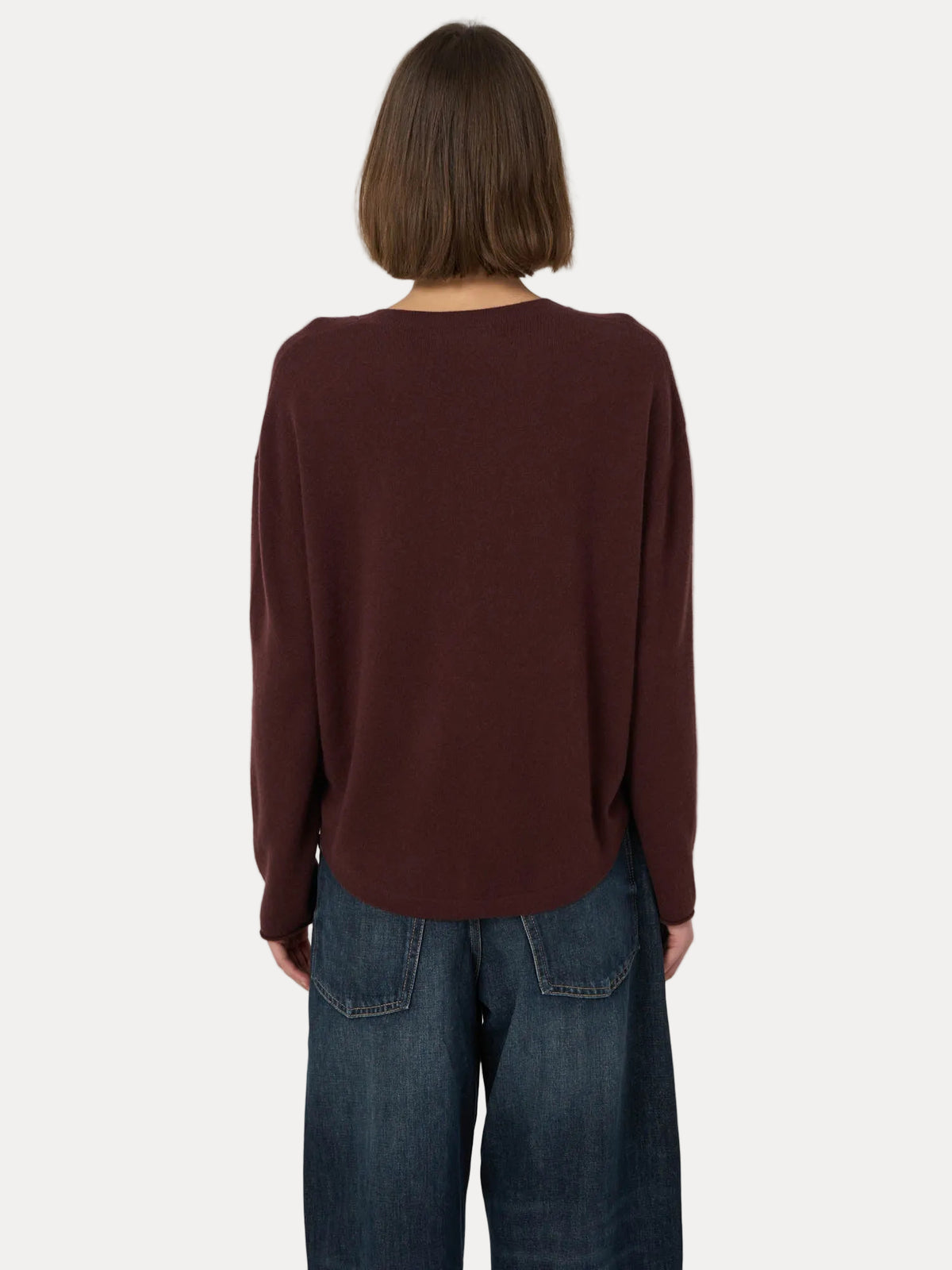 A person viewed from the back wearing a loose, long-sleeve knit top in a deep burgundy-brown color, paired with dark blue jeans, against a plain light gray background.