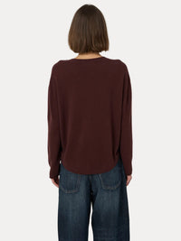 A person viewed from the back wearing a loose, long-sleeve knit top in a deep burgundy-brown color, paired with dark blue jeans, against a plain light gray background.