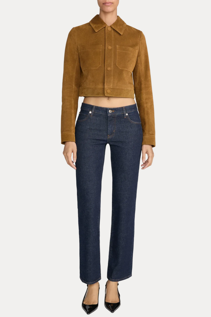 A model wearing a cropped tan suede jacket and dark blue straight-leg jeans, standing against a light gray studio background.
