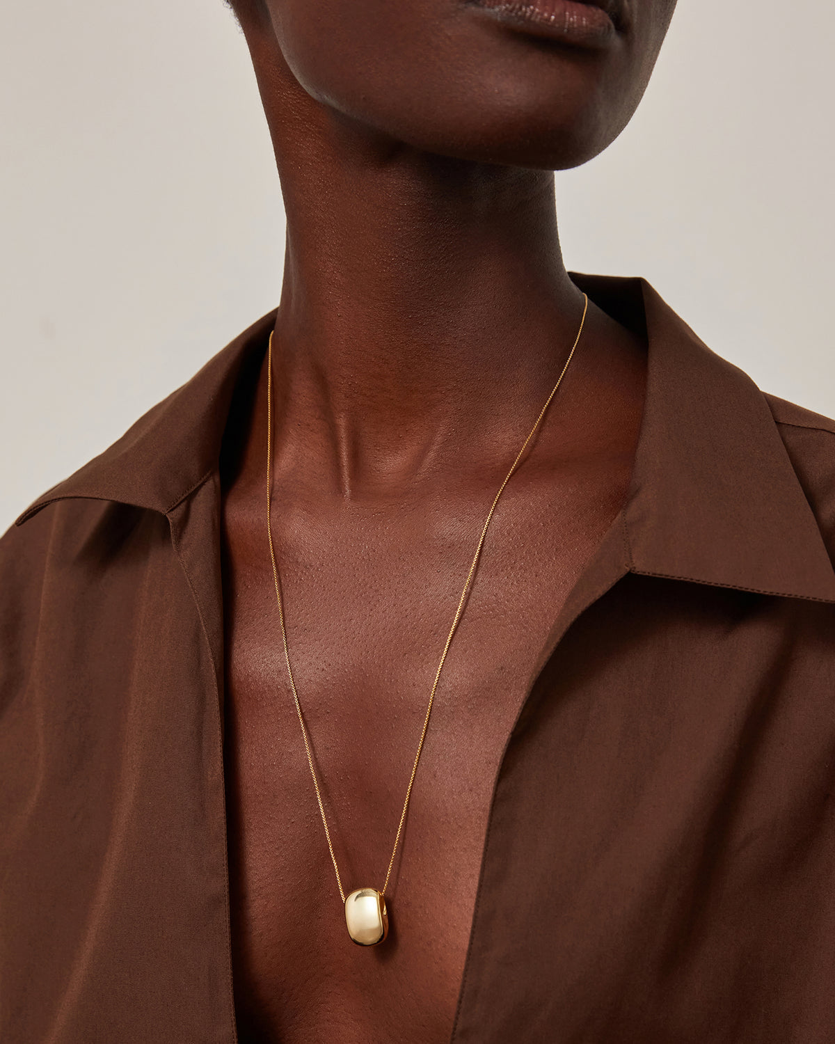 A close-up of a model wearing a delicate gold chain necklace with a small, rounded, cream-colored stone pendant, styled with a brown collared shirt against a neutral light background.