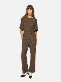 A woman modeling a matching brown short-sleeve top and wide-leg trousers set with a tied waist sash, photographed against a clean light gray studio background.