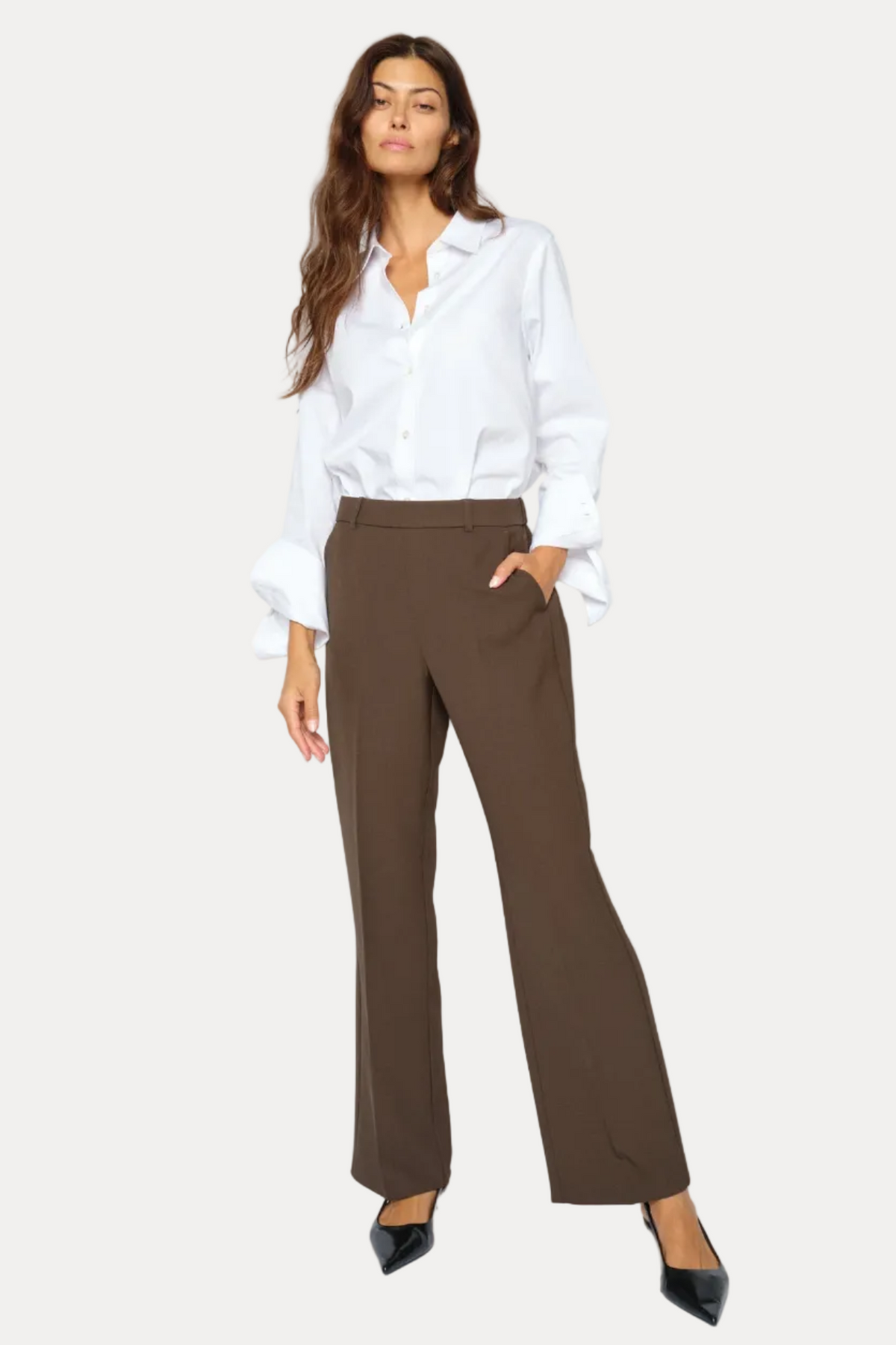 A woman modeling a pair of dark brown wide-leg trousers, paired with a white button-down shirt and black pointed-toe heels, against a plain white studio background.