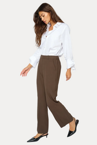 A woman modeling a pair of high-waisted, wide-leg brown trousers paired with a white button-down shirt and black slingback heels, set against a plain white studio background.