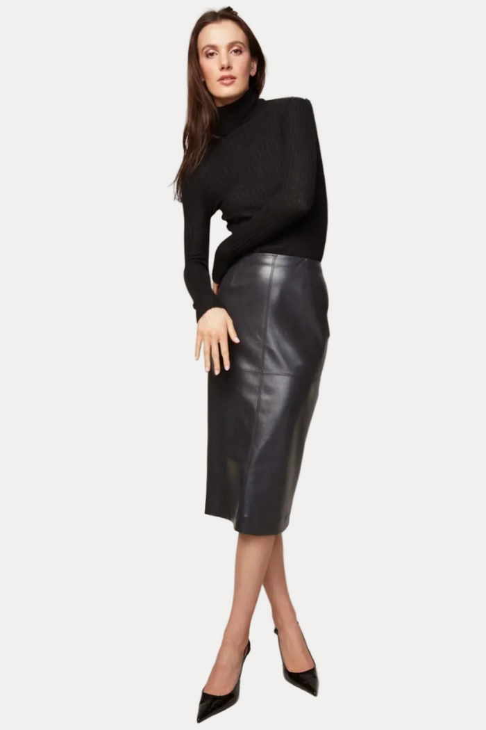 A woman modeling a black ribbed turtleneck sweater and a black leather pencil skirt, paired with black pointed-toe heels, against a solid light gray studio background.