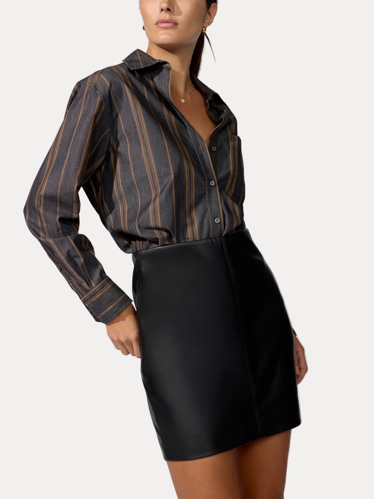 A model wearing a dark gray and black vertically striped button-down shirt tucked into a black faux-leather mini skirt, photographed against a neutral light gray studio background.