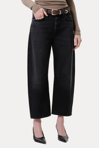 A model wearing black wide-leg cropped jeans with a brown belt, paired with a beige ribbed long-sleeve top and black pointed-toe heels, photographed against a light gray studio background.