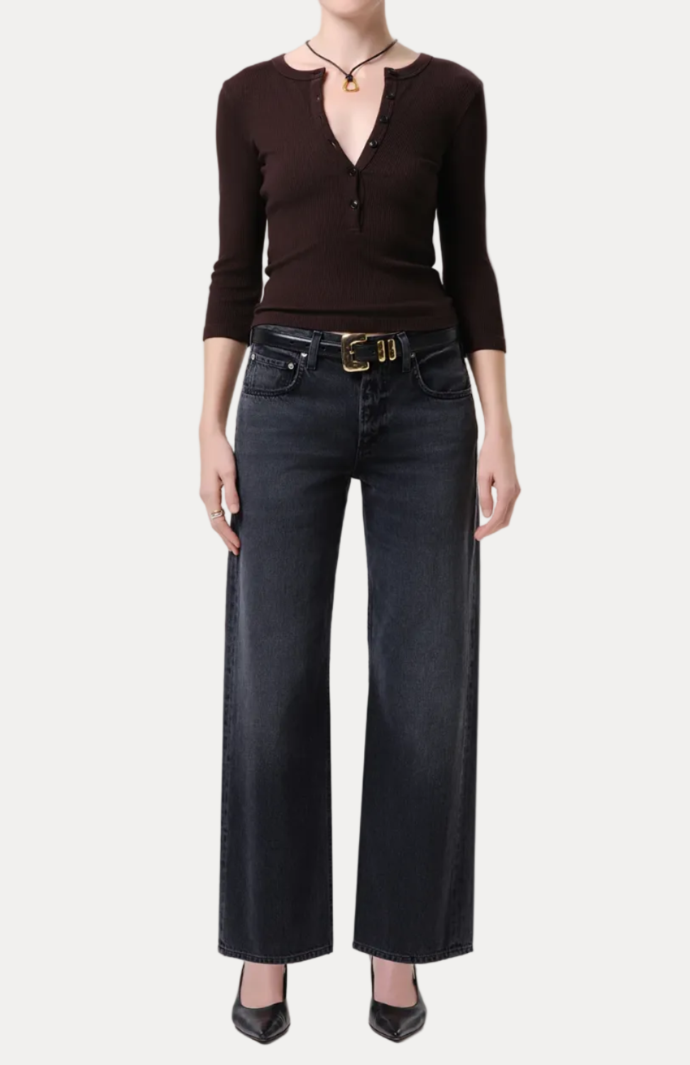 A model wearing a dark brown long-sleeve knit top with a split V-neckline and dark wide-leg jeans with a gold-buckled belt, standing against a light gray studio background.