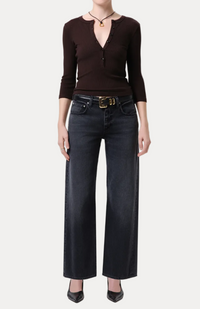 A model wearing a dark brown long-sleeve knit top with a split V-neckline and dark wide-leg jeans with a gold-buckled belt, standing against a light gray studio background.
