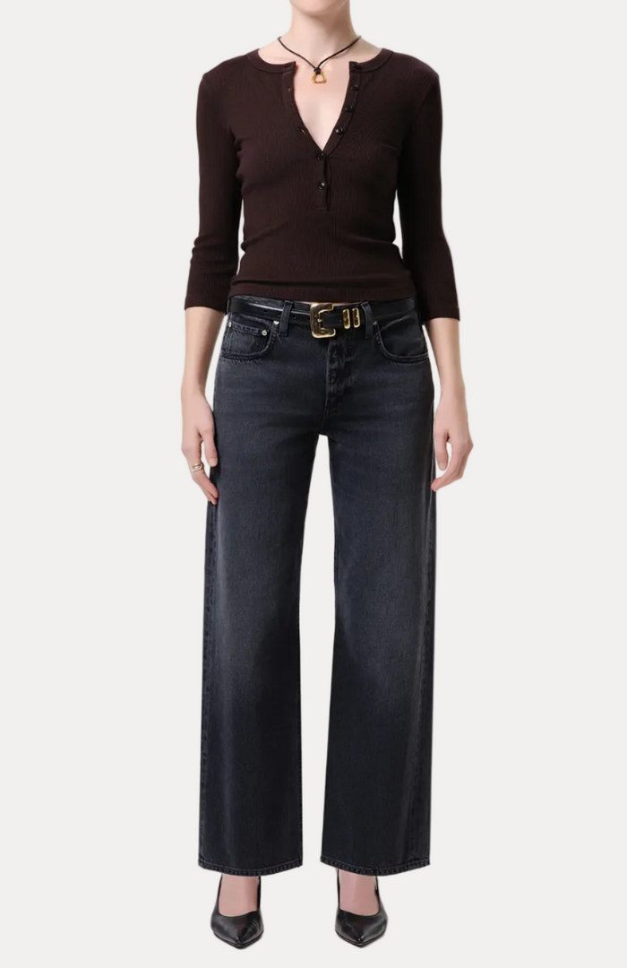 A model wearing a dark brown long-sleeve knit top with a split V-neckline and dark wide-leg jeans with a gold-buckled belt, standing against a light gray studio background.
