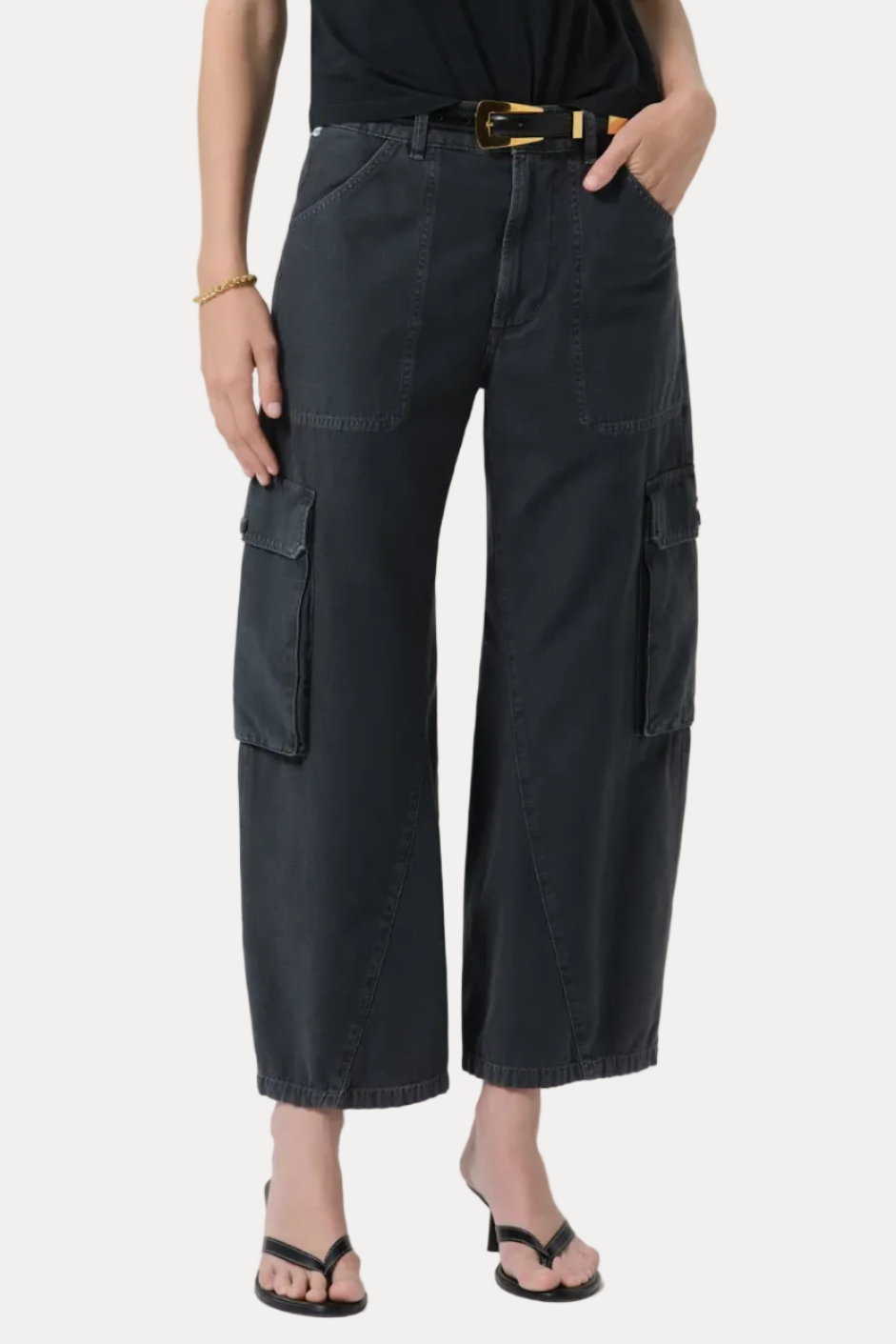 A model wearing dark charcoal gray cropped cargo pants with a black top and black sandals, standing against a light gray studio background.