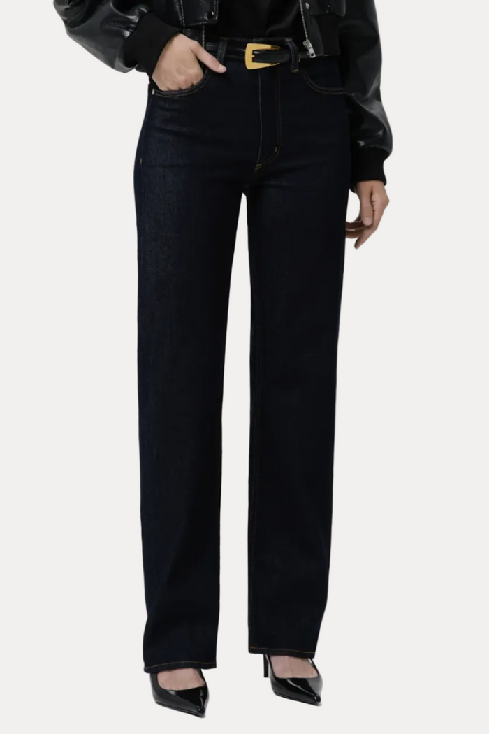 A model wearing high-waisted, straight-leg dark wash jeans with a gold-tone belt buckle, styled with a black leather jacket, black top, and black pointed-toe heels against a light gray studio background.