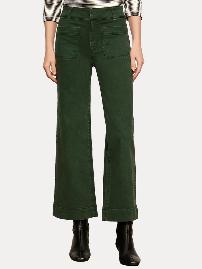 A model is shown from the waist down wearing dark green wide-leg cropped jeans with cuffed hems, paired with a striped top and black ankle boots, against a plain off-white wall and light wood floor.