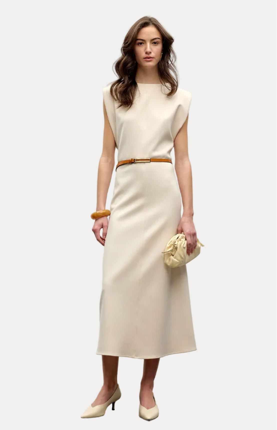 A woman models a sleeveless cream-colored midi dress with a thin tan belt at the waist, holding a matching cream clutch purse and wearing cream heels against a plain light gray studio background.