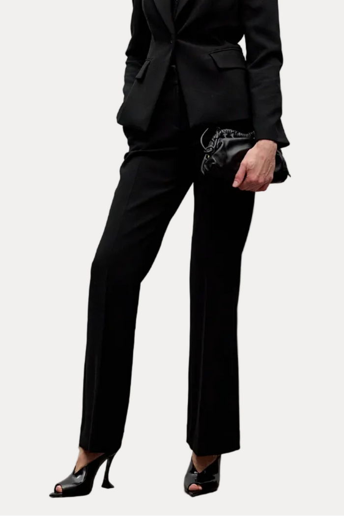 A blonde model wearing a black tailored blazer and matching trousers, styled with black high-heeled sandals and a small black clutch, standing against a minimalist light gray wall.