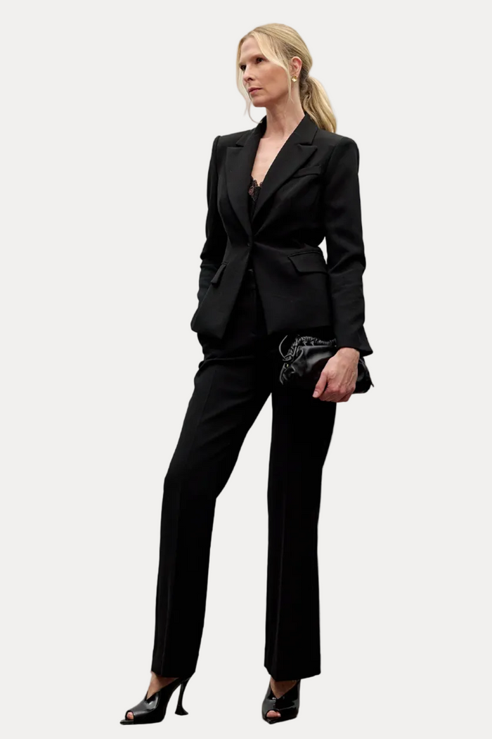 A blonde model wearing a black tailored blazer and matching trousers, styled with black high-heeled sandals and a small black clutch, standing against a minimalist light gray wall.