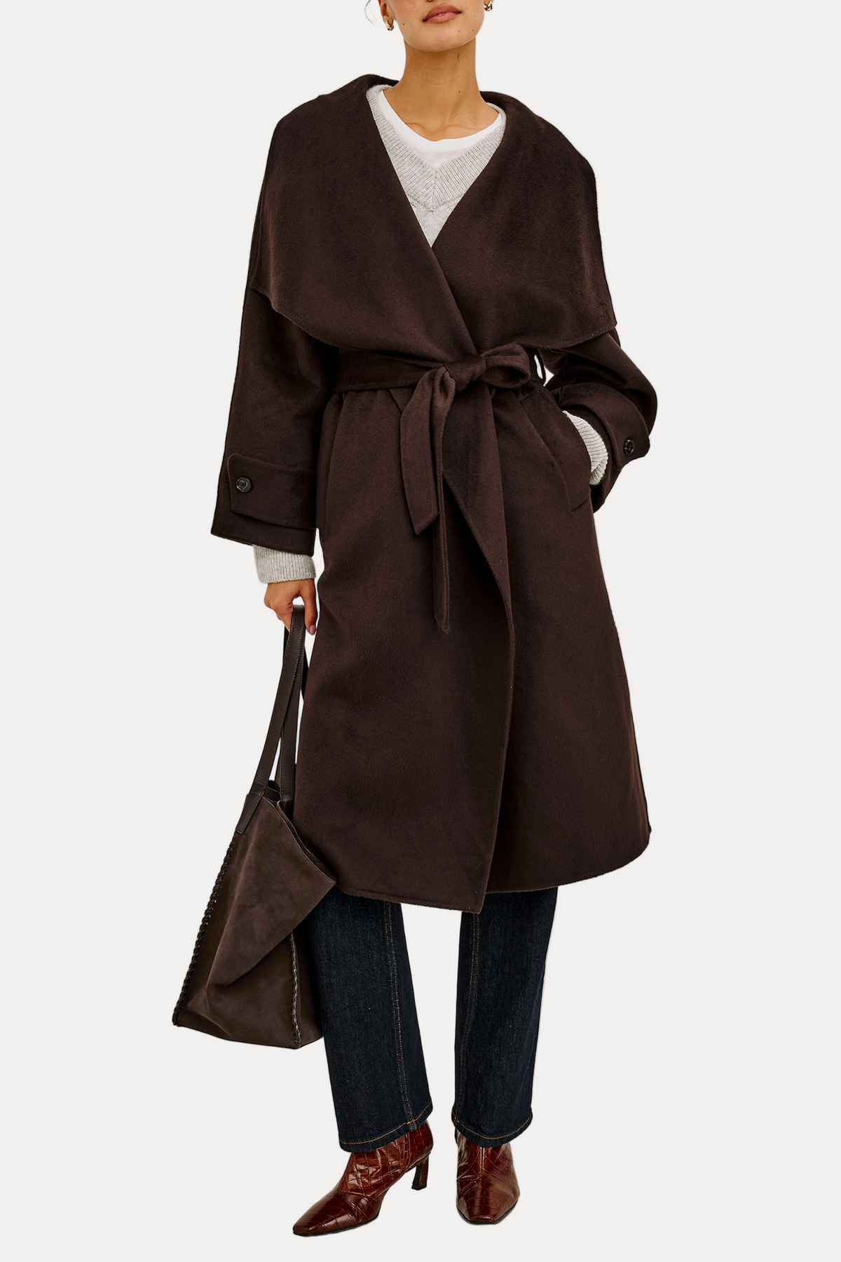 A model wearing a dark brown wrap coat with a tie belt, paired with dark blue jeans, brown pointed-toe boots, and holding a matching dark brown handbag against a plain white background.