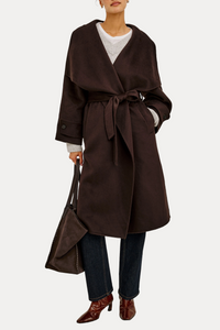 A model wearing a dark brown wrap coat with a tie belt, paired with dark blue jeans, brown pointed-toe boots, and holding a matching dark brown handbag against a plain white background.