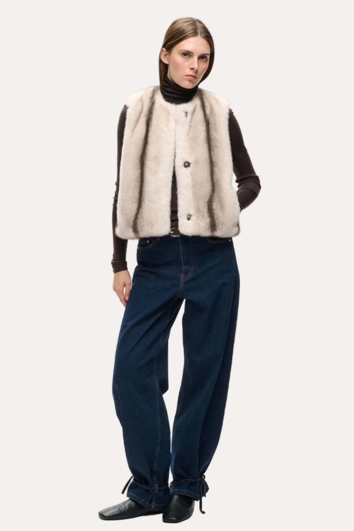 A model stands against a light gray studio background wearing a beige and brown striped faux-fur vest over a black turtleneck, paired with dark blue wide-leg jeans and black shoes.