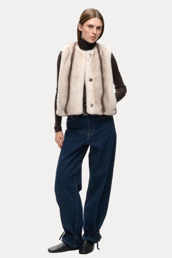 A model stands against a light gray studio background wearing a beige and brown striped faux-fur vest over a black turtleneck, paired with dark blue wide-leg jeans and black shoes.