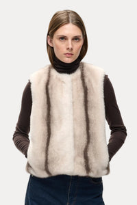 A model wearing a cream-colored shaggy faux fur vest over a dark brown turtleneck sweater and dark blue jeans, shot against a plain white studio background.