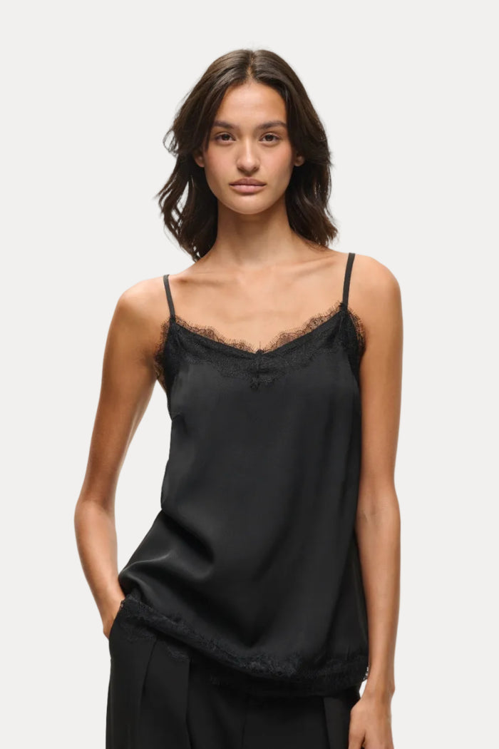 A woman modeling a black satin camisole top with delicate lace trim along the neckline and hem, paired with black trousers, against a plain light gray studio background.