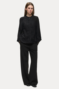 A woman modeling a black long-sleeve button-front blouse with lace detailing on the front, paired with black wide-leg trousers, against a plain light gray studio background.
