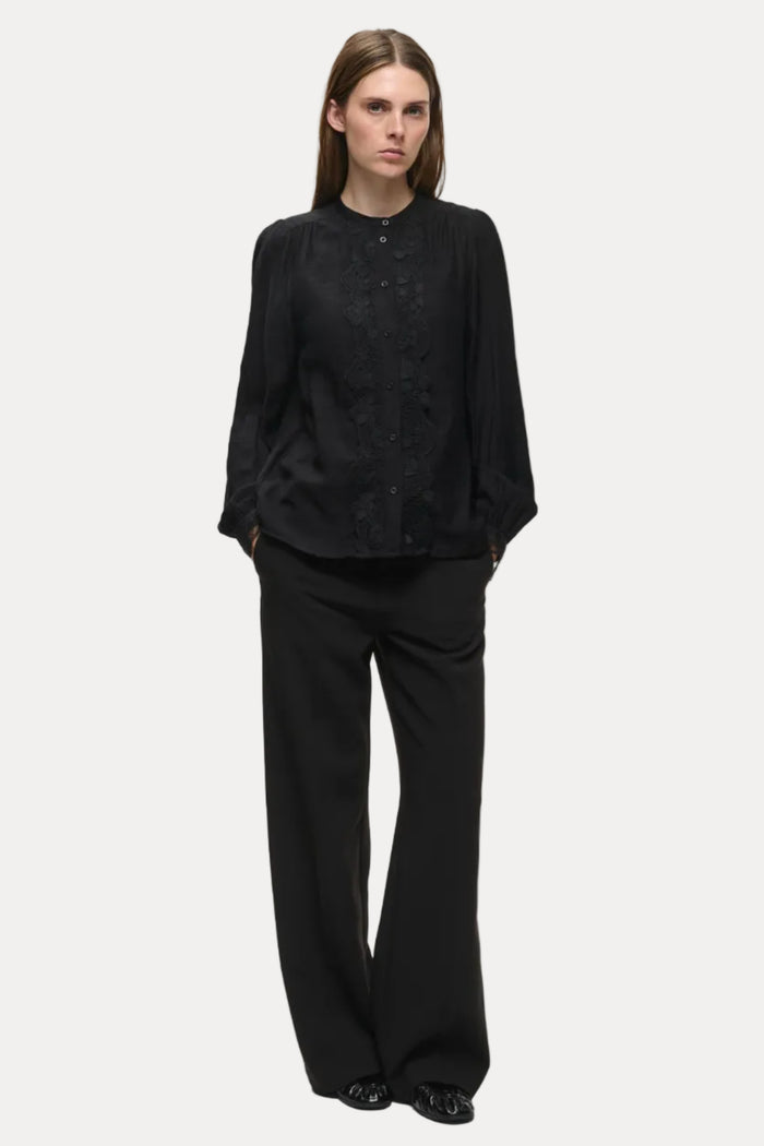 A woman modeling a black long-sleeve button-front blouse with lace detailing on the front, paired with black wide-leg trousers, against a plain light gray studio background.