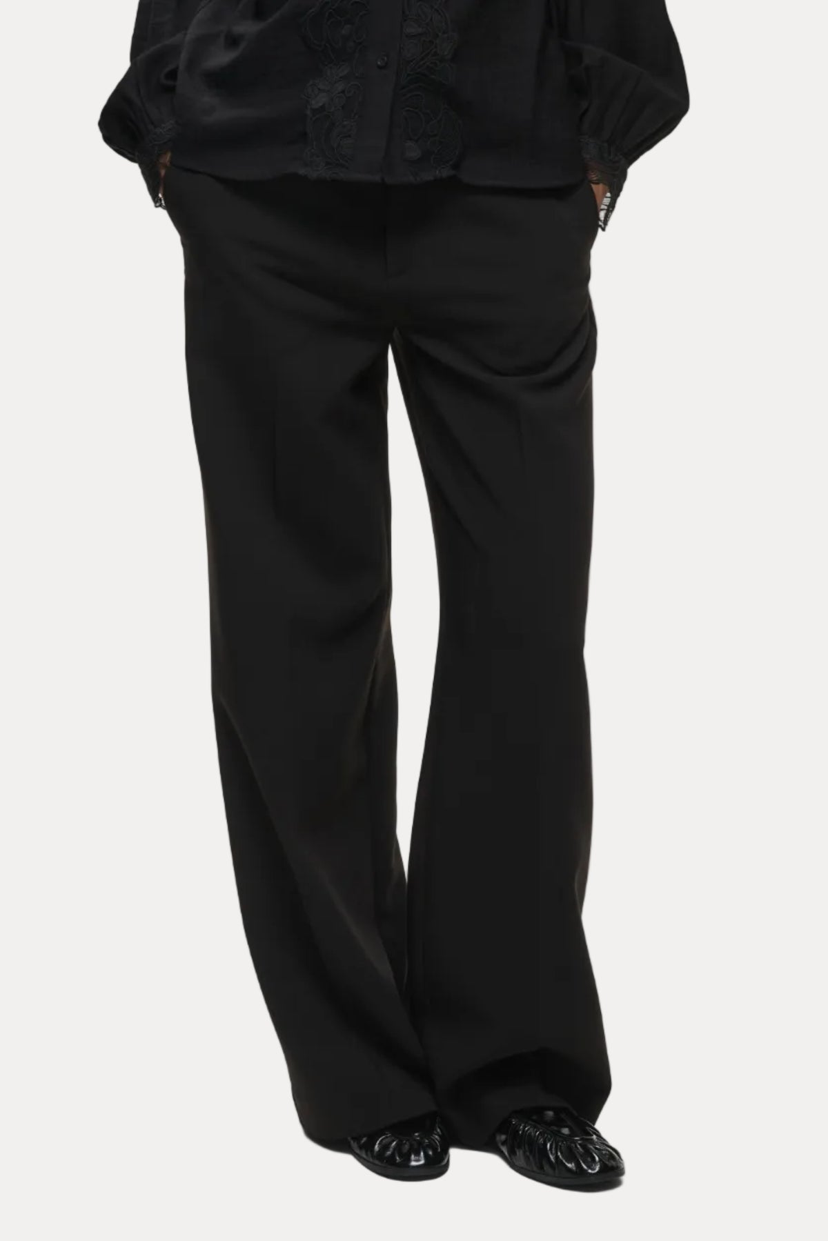 A person wearing black wide-leg trousers with a black blouse and black shoes, photographed against a plain light gray studio background.