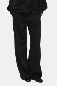 A person wearing black wide-leg trousers with a black blouse and black shoes, photographed against a plain light gray studio background.