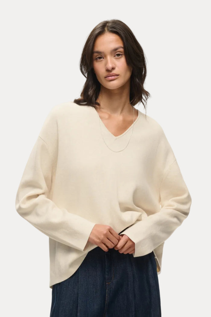A woman modeling a cream-colored, long-sleeve knit sweater with a V-neck, paired with dark blue wide-leg jeans, against a plain white studio background.