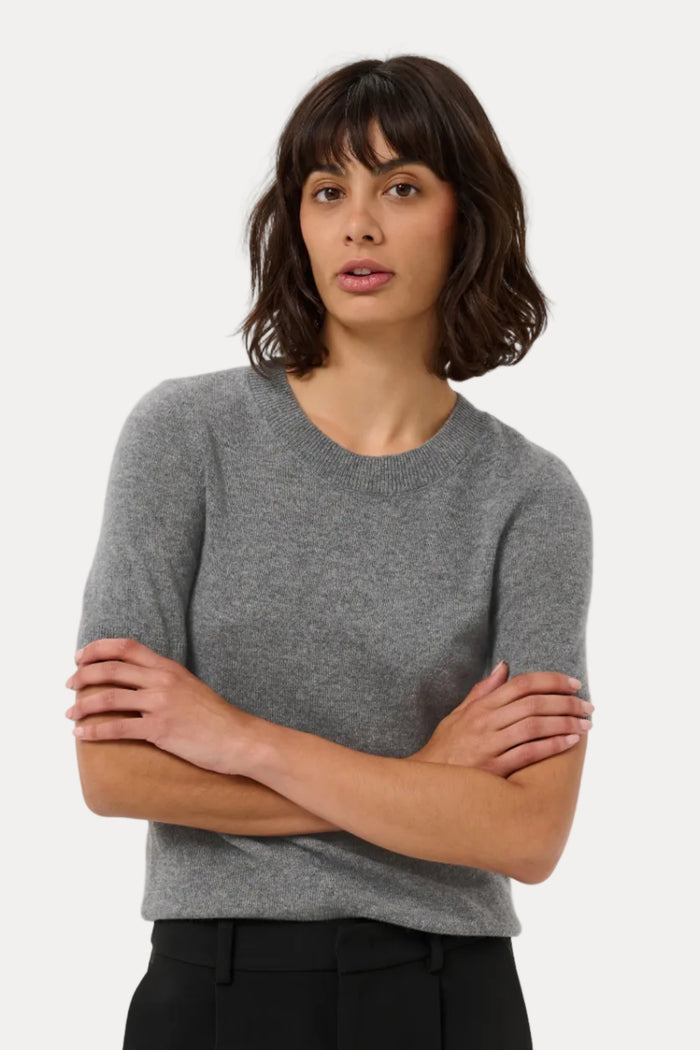 A woman modeling a short-sleeve crewneck knit sweater in heather gray, paired with black trousers, against a plain light gray studio background.