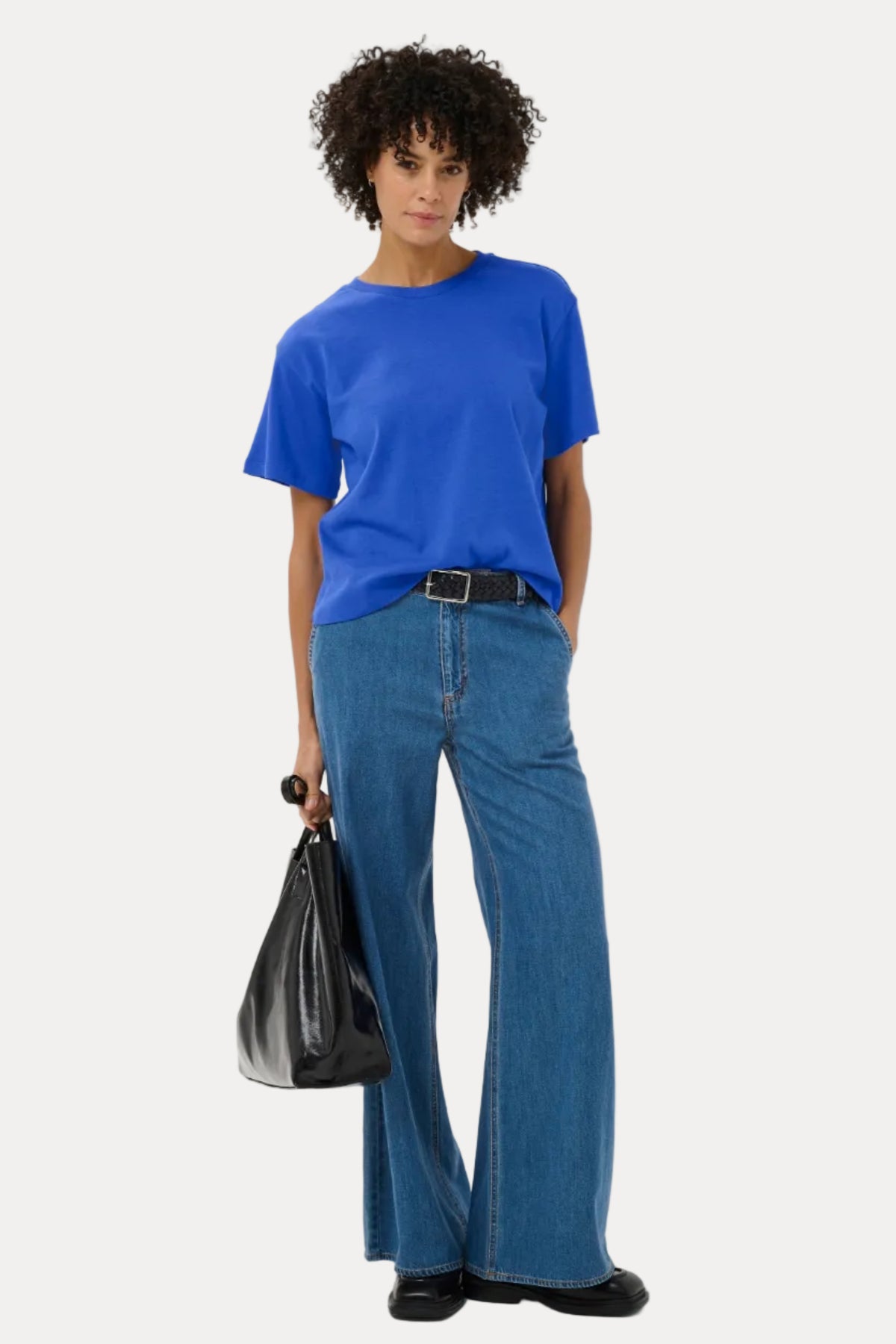 A model stands against a light gray studio background wearing a bright blue short-sleeve crewneck t-shirt, wide-leg blue jeans, a black belt, black shoes, and holding a black leather handbag.