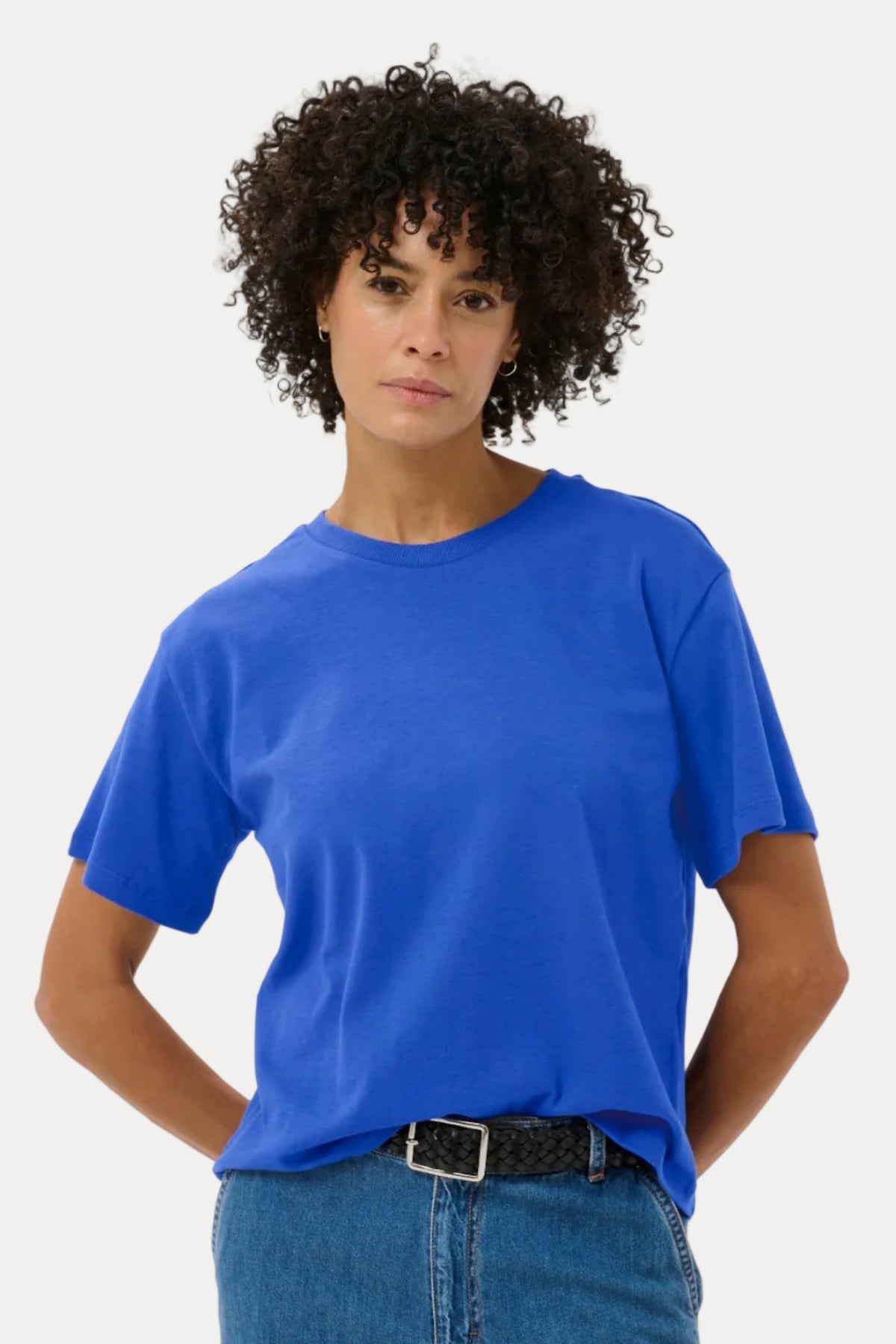A model with curly dark hair wearing a bright royal blue short-sleeve crewneck t-shirt and blue jeans with a black belt, standing against a light gray studio background.