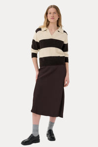 A model stands against a light gray studio background wearing a knit polo-style sweater with wide horizontal stripes in cream and black, paired with a dark brown midi skirt, gray socks, and black shoes.