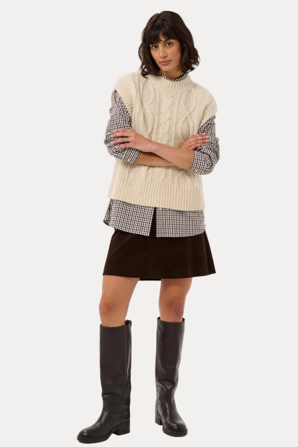 A model stands against a light gray studio background wearing a cream cable-knit sweater vest over a black-and-white gingham shirt, a dark brown mini skirt, and tall black leather boots.