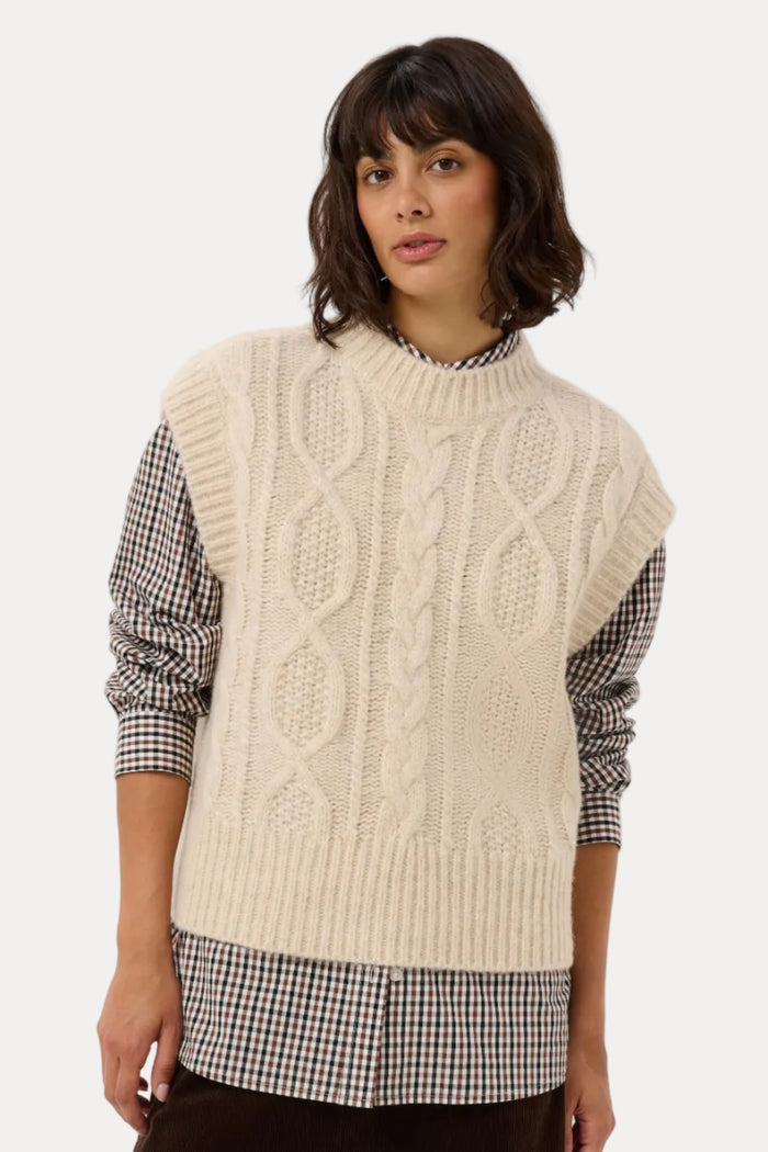 A woman modeling a cream cable-knit mock-neck sweater vest over a black and white gingham shirt and a dark brown skirt, against a light gray studio background.