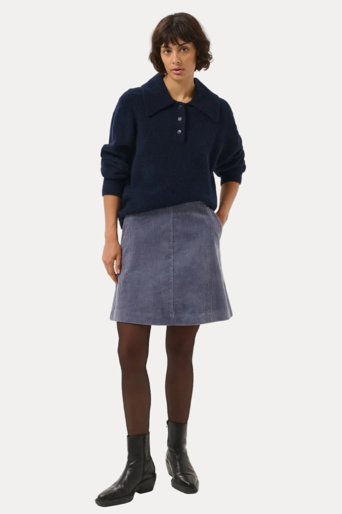 A model stands against a light gray studio background wearing a dark navy long-sleeve polo sweater, a slate blue corduroy A-line skirt, dark brown tights, and black ankle boots.