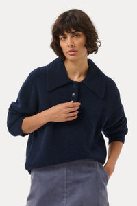 A woman modeling a dark navy blue knit polo sweater paired with a slate blue A-line skirt, photographed against a seamless light gray studio background.
