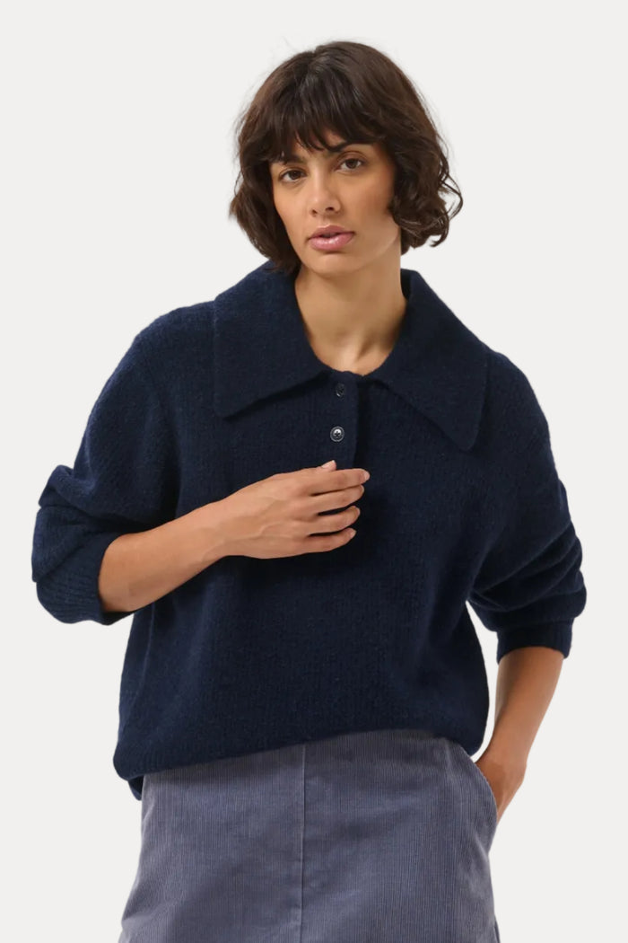 A woman modeling a dark navy blue knit polo sweater paired with a slate blue A-line skirt, photographed against a seamless light gray studio background.