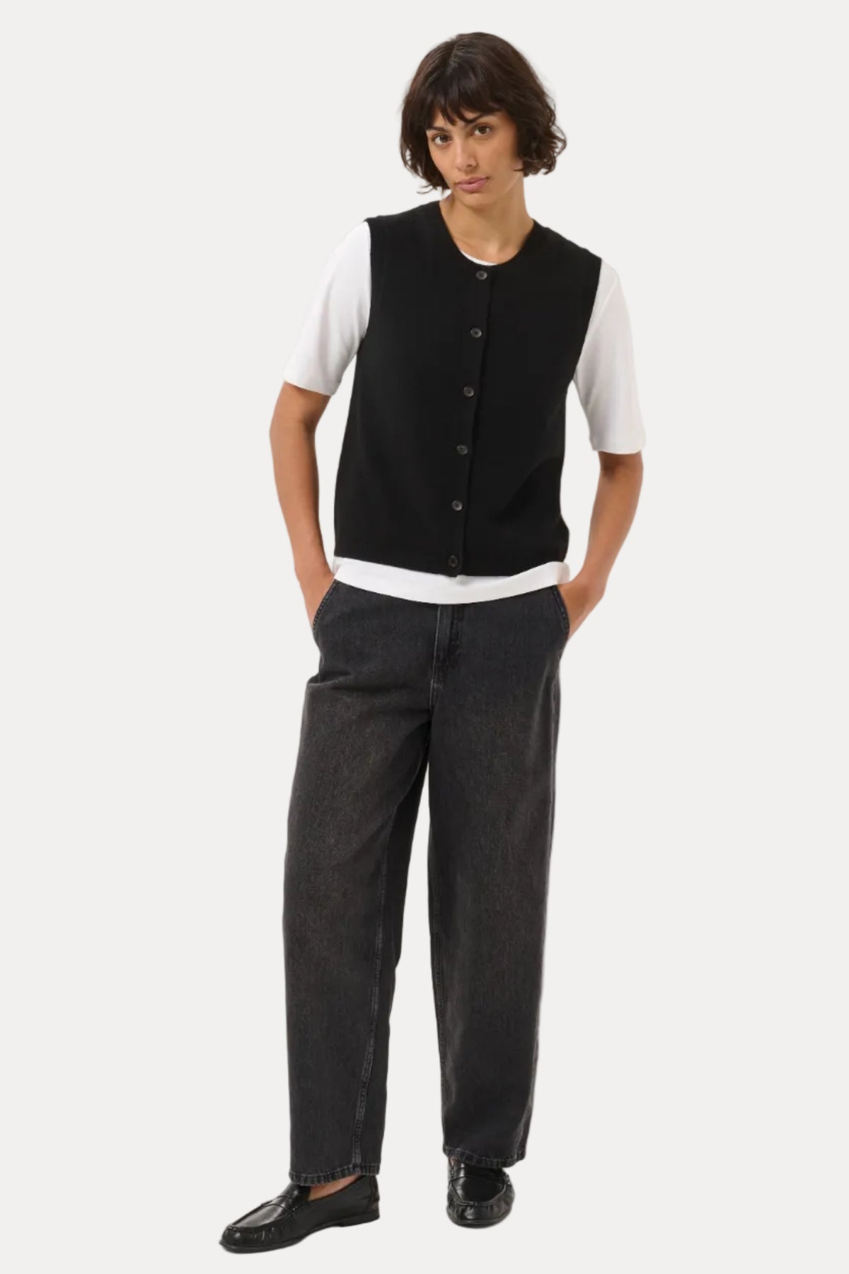 A model stands against a light gray studio background wearing a black sleeveless button-front vest over a white short-sleeve T-shirt, dark charcoal gray wide-leg jeans, and black loafers.